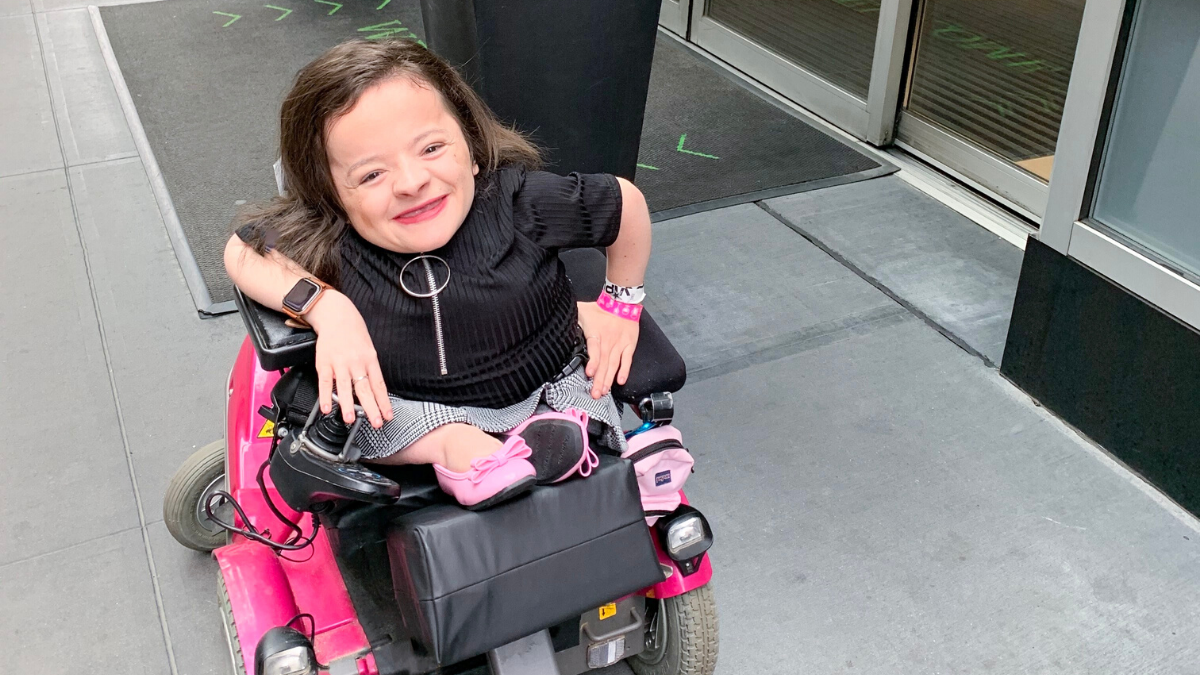 Michaela Davert in pink motorized wheelchair, black shirt, plaid skirt, and pink flats